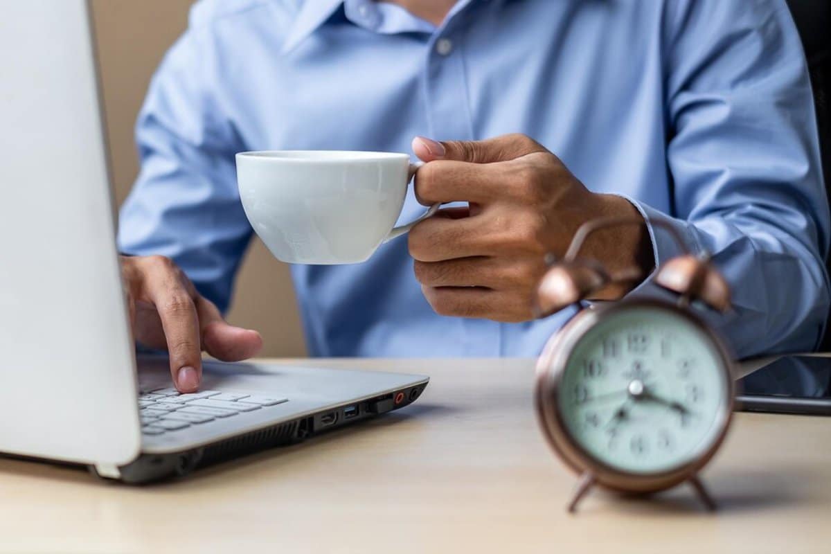 Man in blue shirt holding a coffee cup while using a laptop, with a clock in the foreground.
