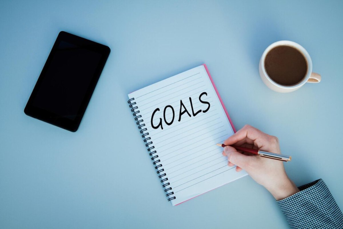 Hand writing GOALS in a notebook, with a smartphone and coffee mug on a blue desk.