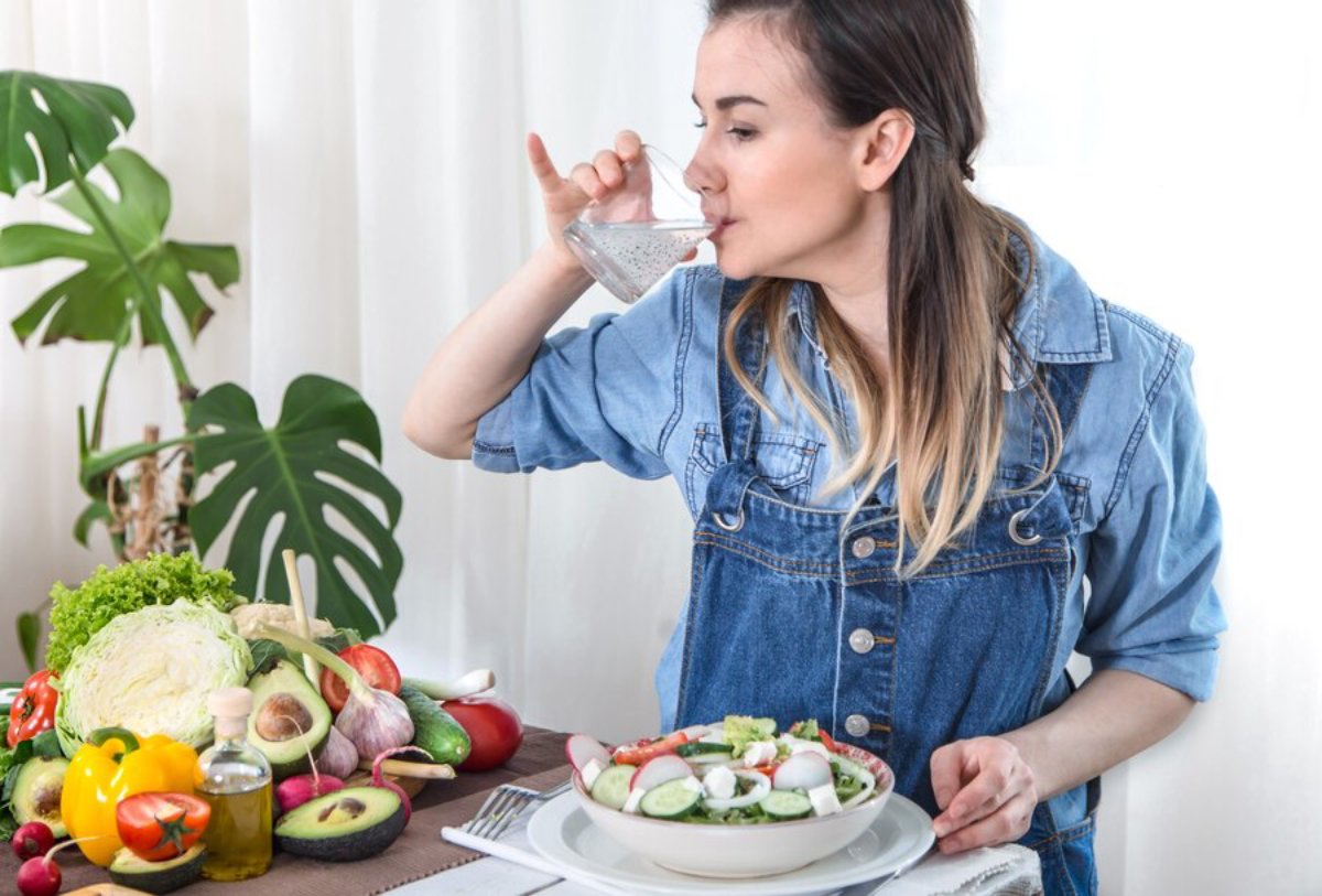 Young woman drinking water at a table with a plate of salad and fresh vegetables.