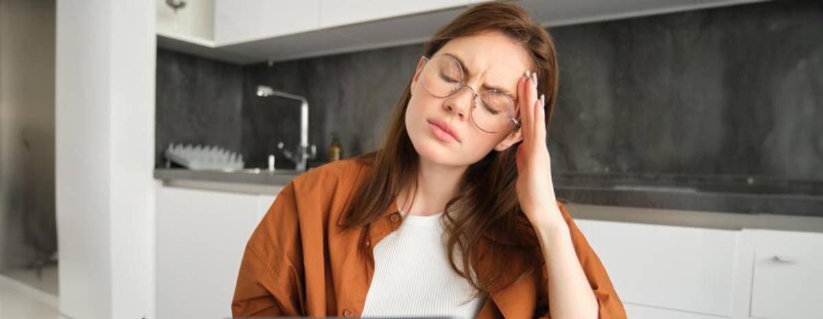 Young woman in glasses looks stressed or fatigued, holding head, in a modern kitchen.