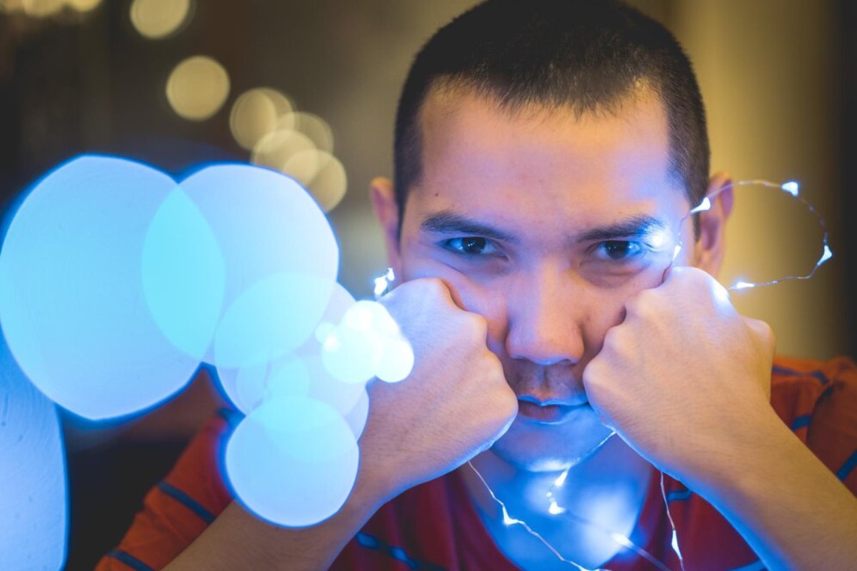 Young man with a serious expression, holding string lights near his face, with blurred lights in the background.