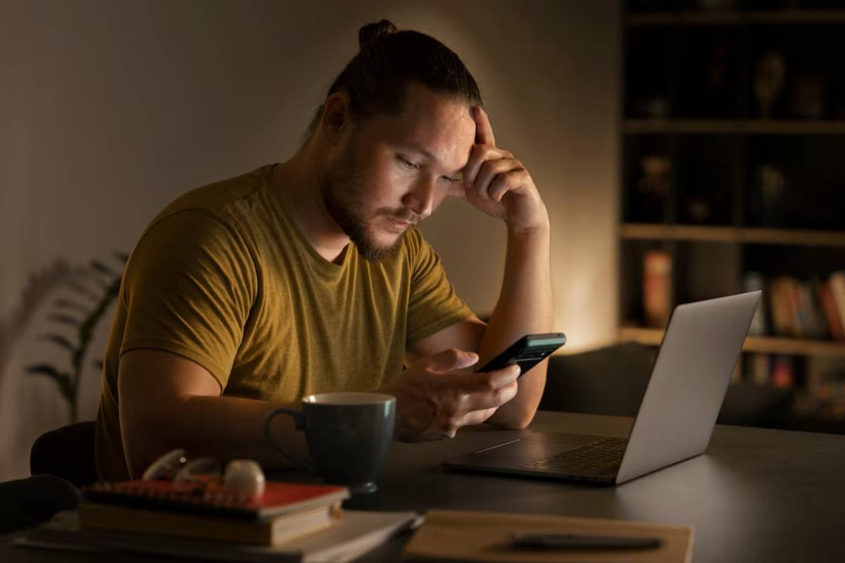 Man looking at his smartphone with concern while sitting at a desk with a laptop and coffee mug.
