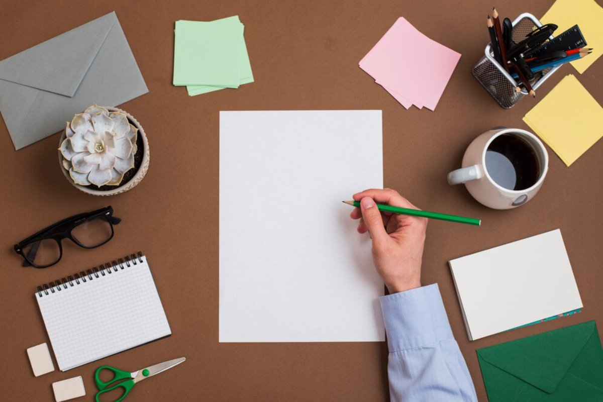 Overhead view of a person's hand writing on a blank sheet with office supplies and coffee around on a brown desk.