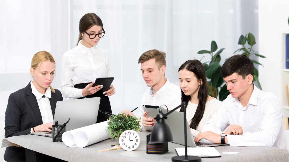 Five professionals collaborating at a modern office desk with computers, tablets, and paperwork.