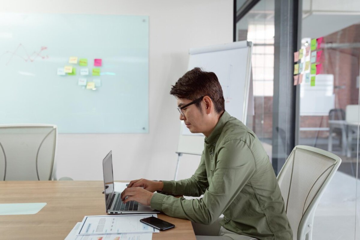 Man in glasses working on a laptop in a modern office space with a whiteboard and sticky notes in the background.