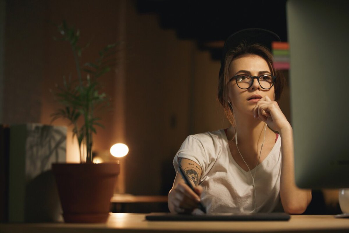 Young woman with glasses working at a computer in a dimly lit room at night.