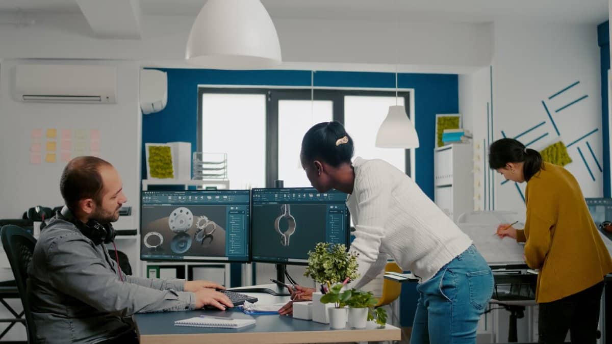 Three professionals work in a brightly colored office with one using a computer and two discussing over blueprints.