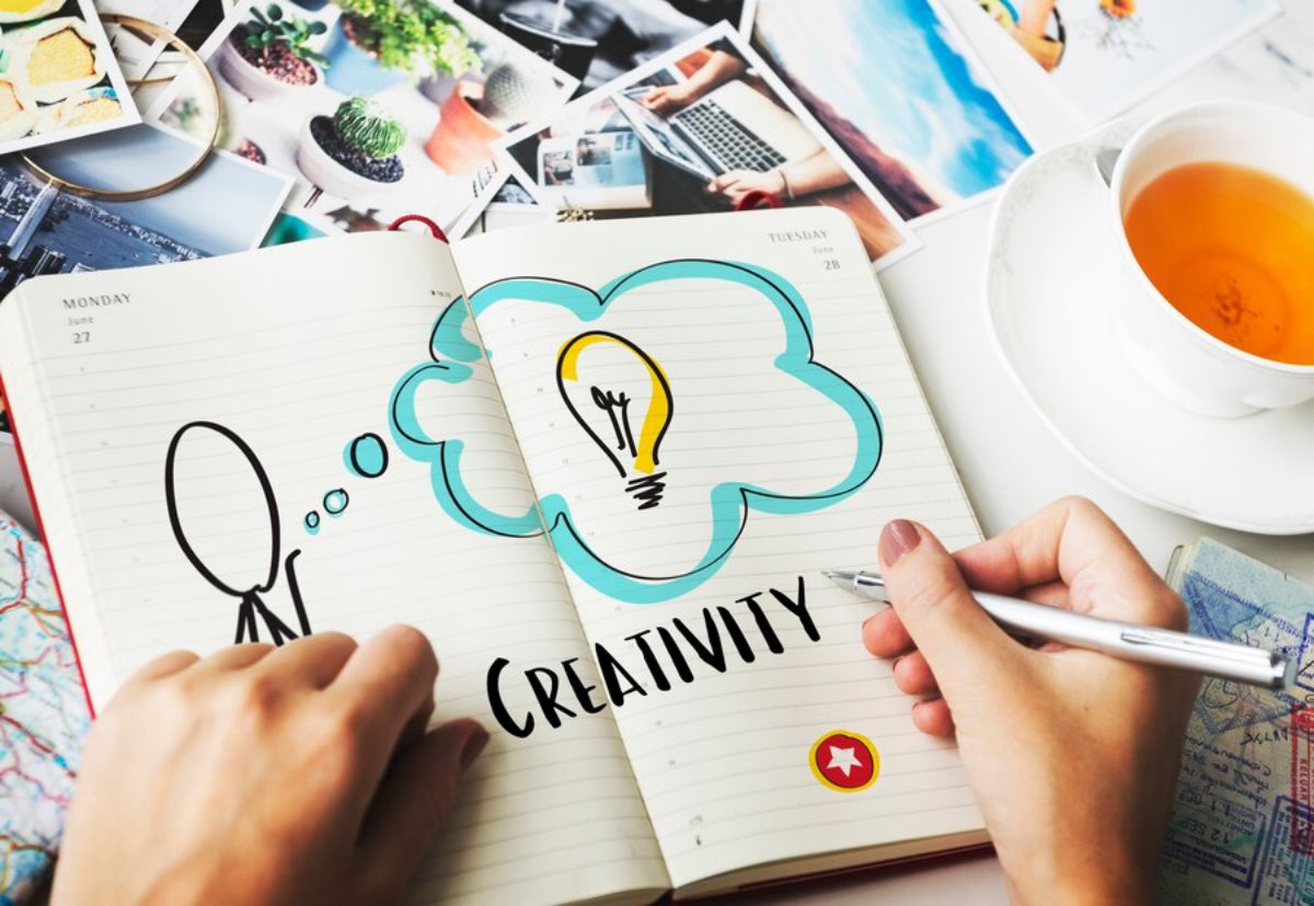 A person doodles a light bulb inside a cloud shape labeled CREATIVITY in their notebook surrounded by diverse images and a cup of tea.