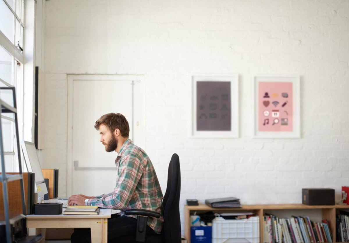 Man in a plaid shirt working at a computer in a bright office with framed artwork on the wall.