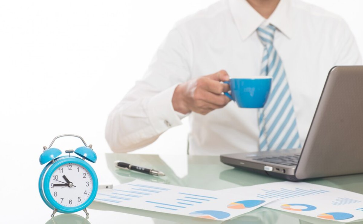 Businessman at desk with laptop, charts, and blue alarm clock, holding a blue coffee mug.