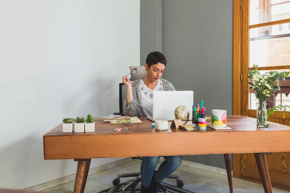 Woman working at a wooden desk with a laptop, surrounded by stationery and plants, in a well-lit room.