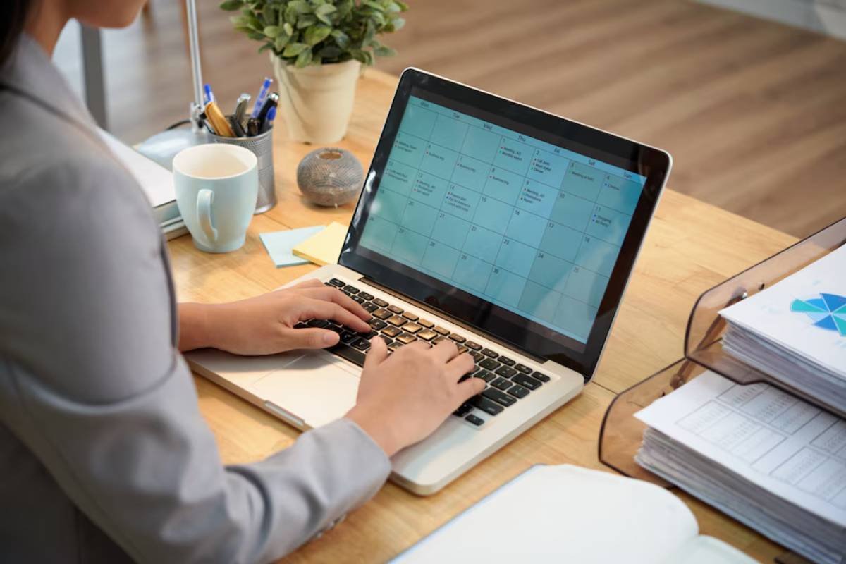 Professional woman working on a laptop displaying a calendar, surrounded by office supplies and a coffee mug.