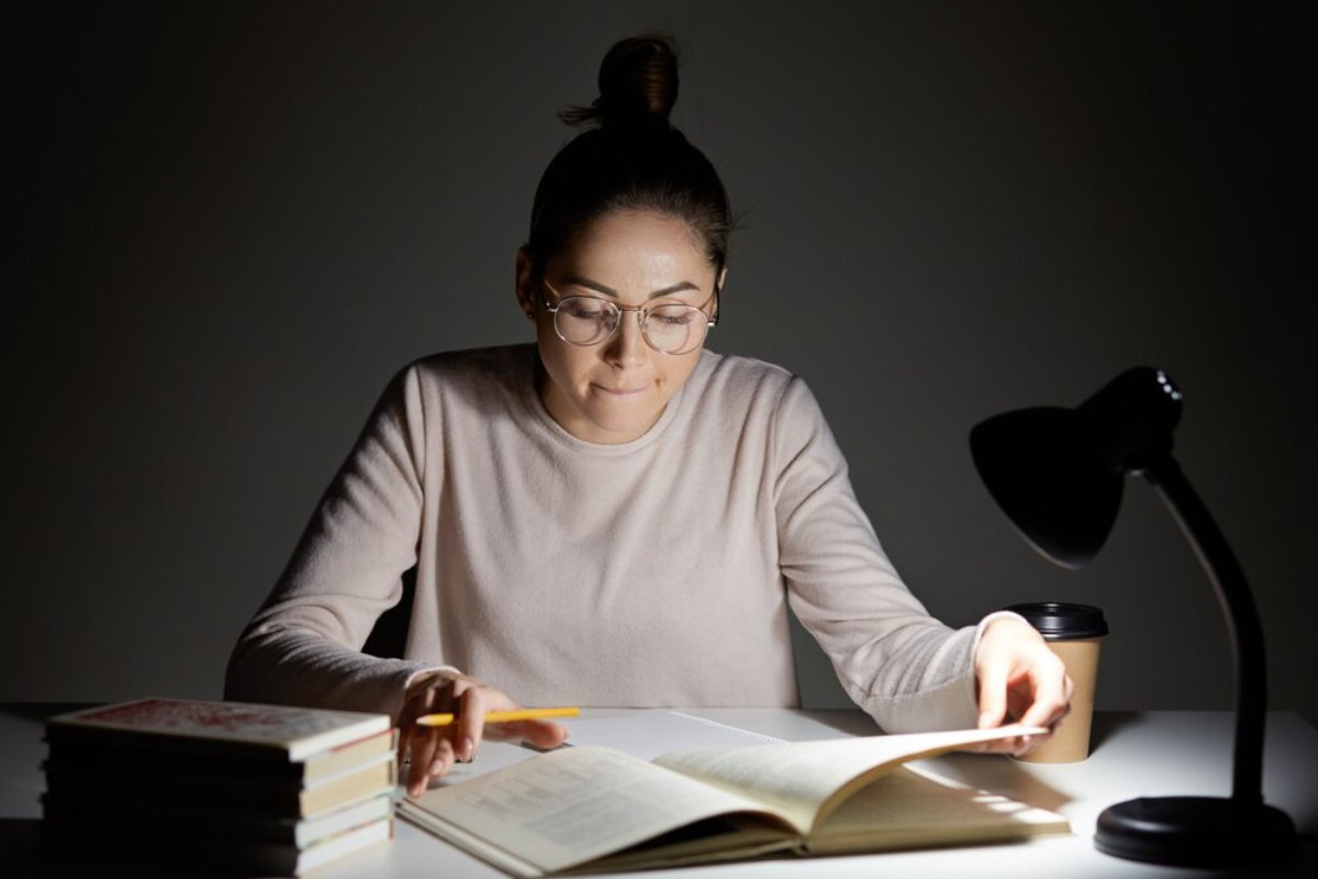 A person in a light sweater studies at a table, illuminated by a desk lamp, surrounded by books and a coffee cup.