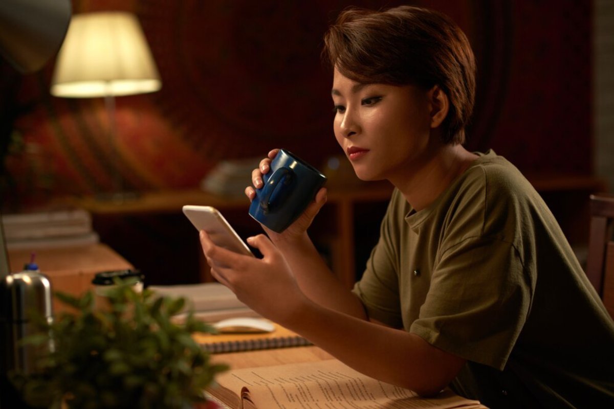 A woman sits at a table, looking at her smartphone while holding a blue mug, in a warmly lit room.
