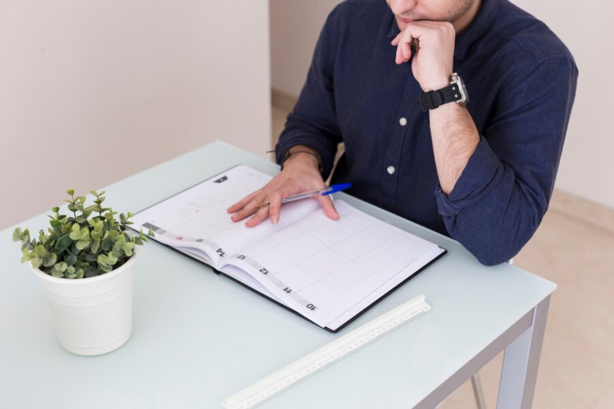 A person in a navy shirt thoughtfully reviews a planner, pen in hand, with a small potted plant on a glass desk.