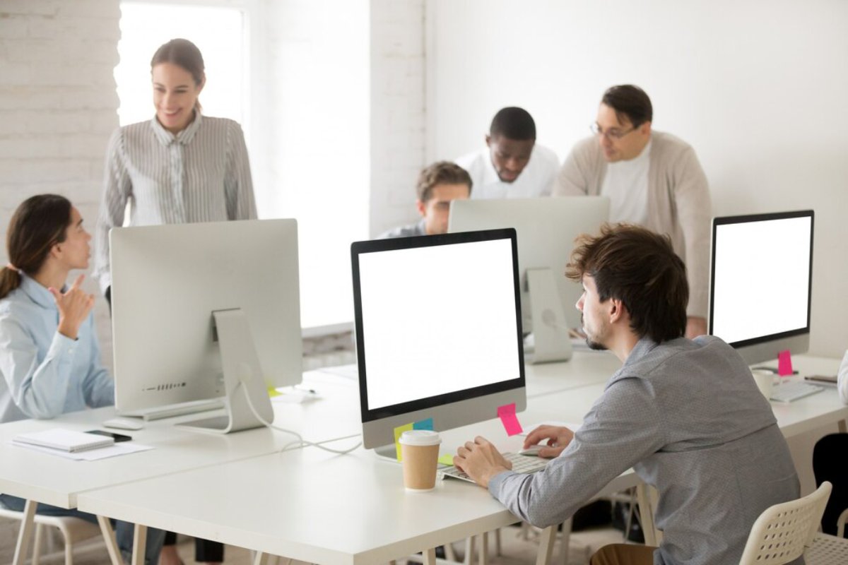 A group of people collaborates in a bright, modern office space, focused on desktop computers with stationery and coffee cups on their desks.