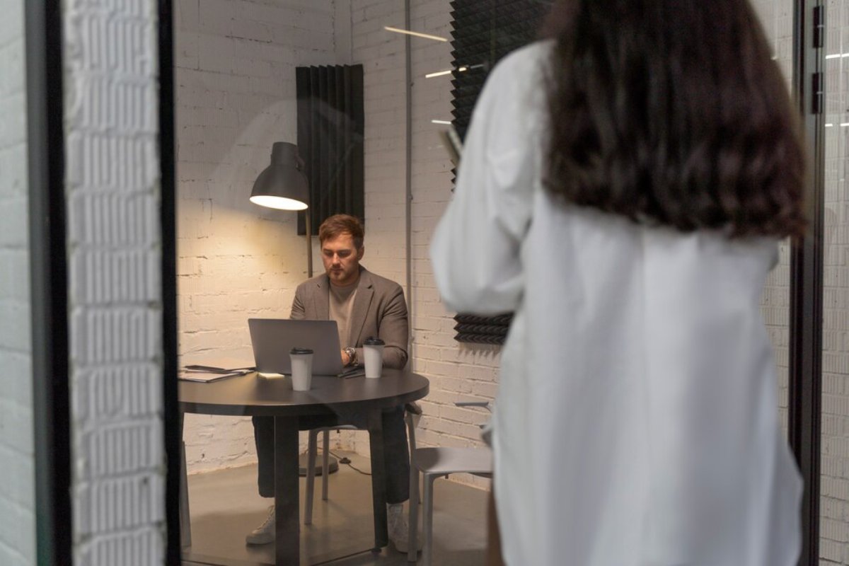 Man working on laptop in modern office space, observed by a woman in white blouse from a blurred foreground.