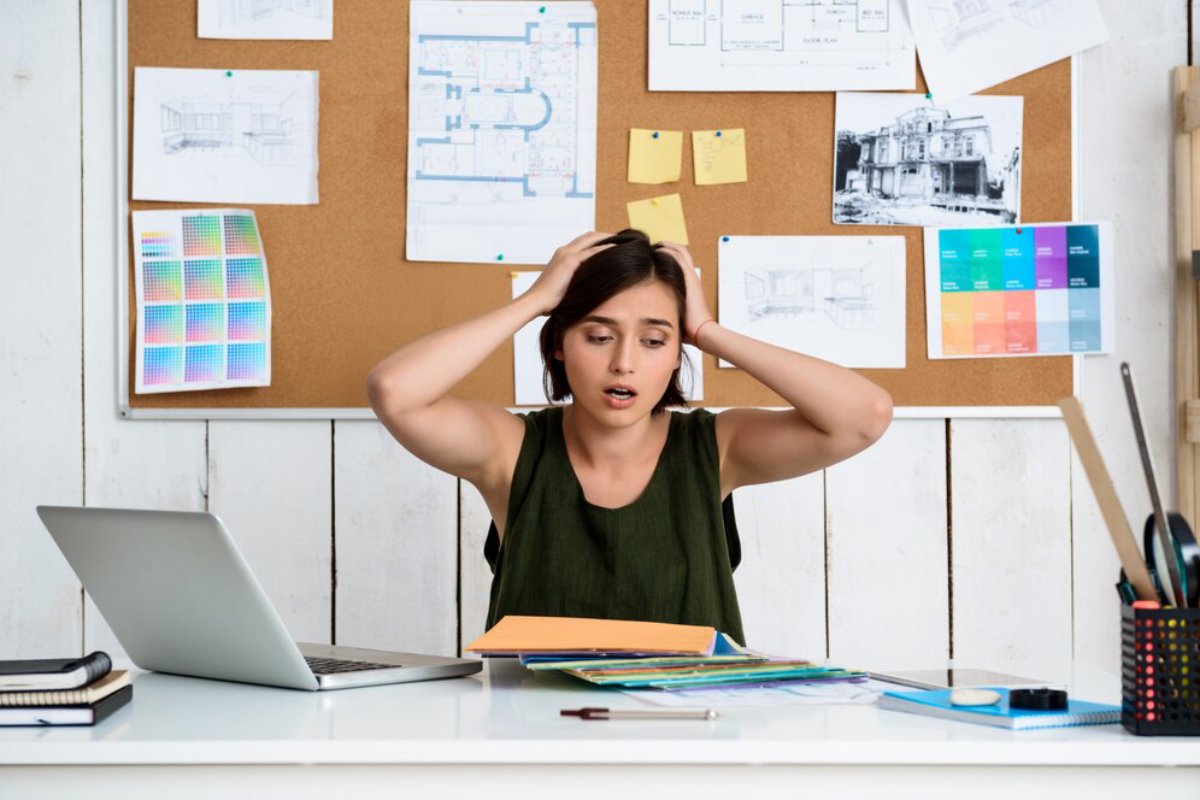 Stressed woman at desk with laptop, surrounded by architectural plans and color swatches.