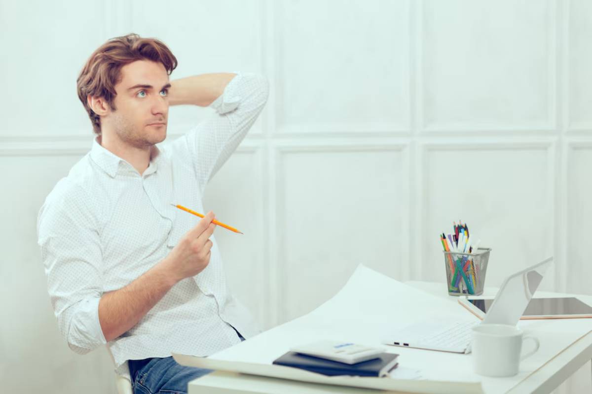 Young man sitting at a desk, looking thoughtful with a pencil in hand, surrounded by papers and a laptop.