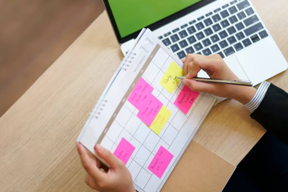 Person planning schedule with sticky notes on a calendar beside an open laptop.