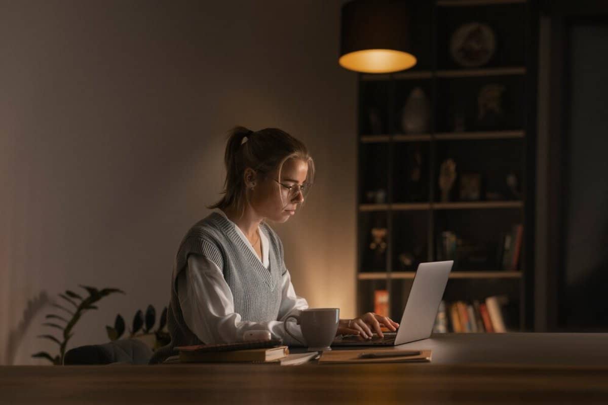 Woman working on laptop in a dimly lit room with a warm lamp overhead.