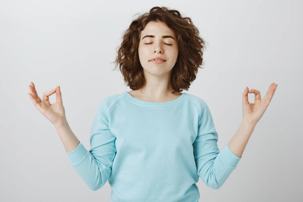 Woman in blue sweater meditating with eyes closed and hands in mudra gesture against a white background.