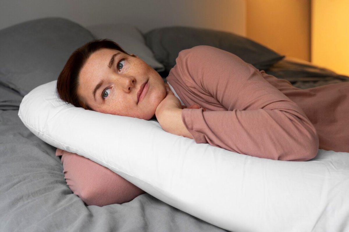 Woman lying on her side in bed, resting her head on a white pillow, looking contemplative.