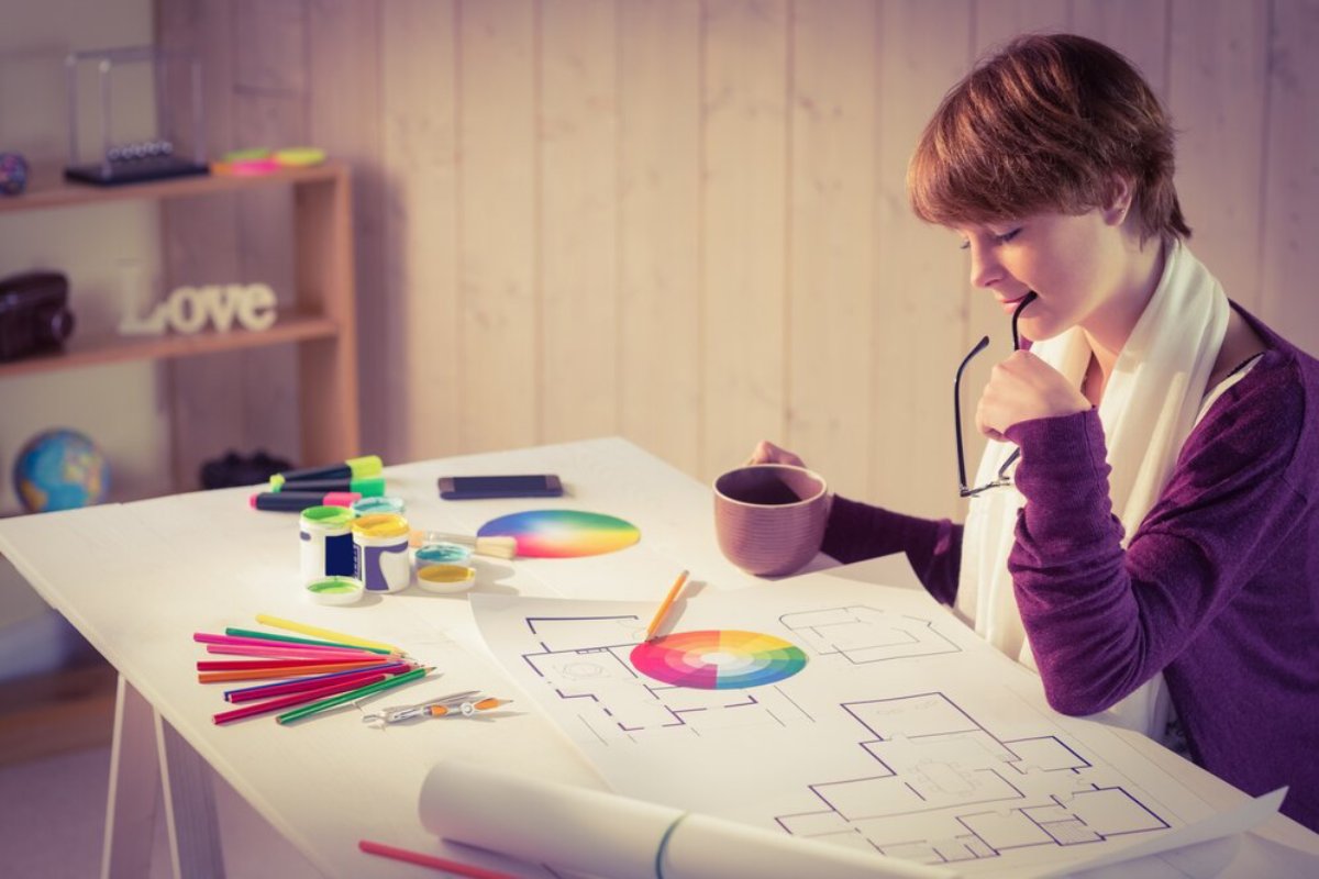 Young woman reviewing architectural drawings at a desk with colored pencils, paint, and a coffee mug.