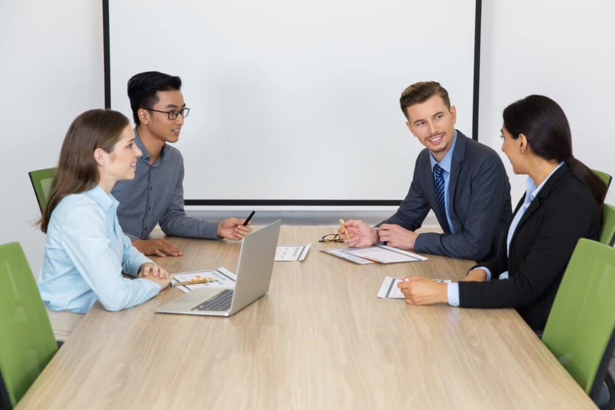Four business professionals happily discussing around a conference table with laptops and documents.