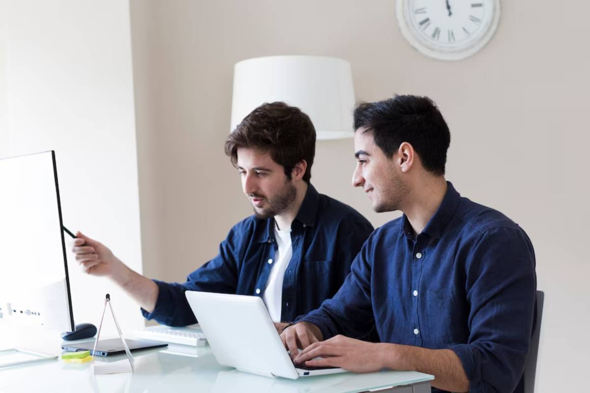 Two men collaborating at a desk with laptops and a computer monitor in an office.