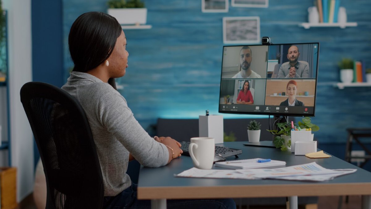 A woman participates in a video conference from an office with four other virtual attendees displayed on her monitor.