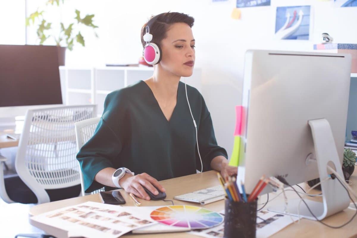 Woman in office using headphones and computer, surrounded by color swatches and office supplies.