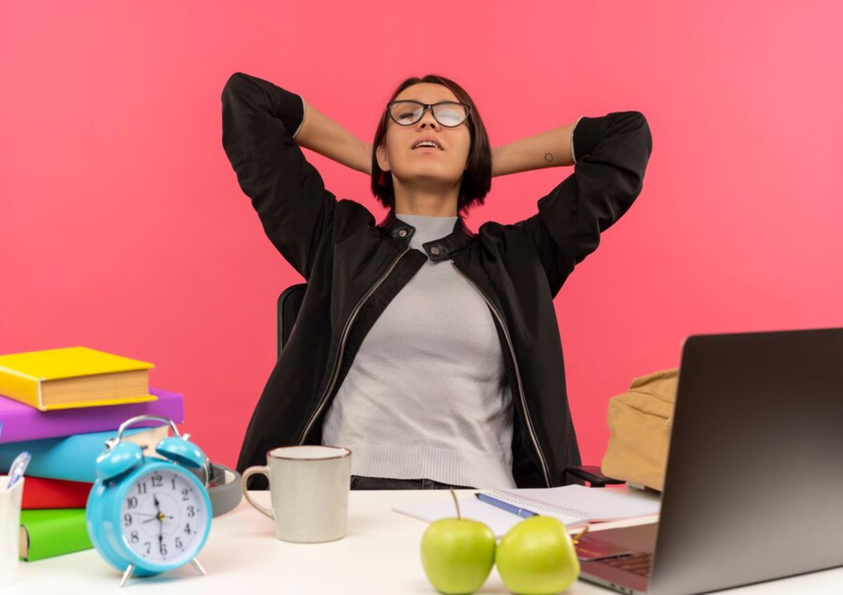 Young woman relaxing with hands behind her head at a desk with books, laptop, and a blue clock, against a pink background.