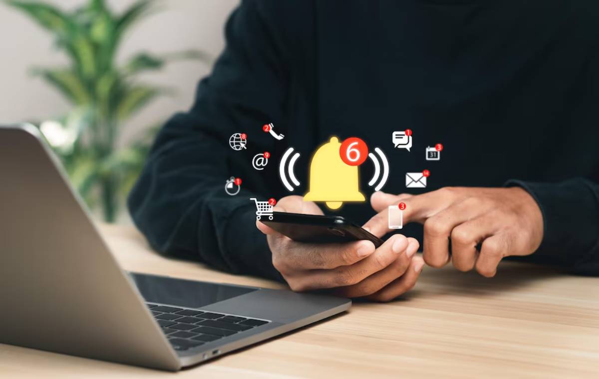 Person using smartphone with notification icons floating above, next to a laptop on a wooden desk.