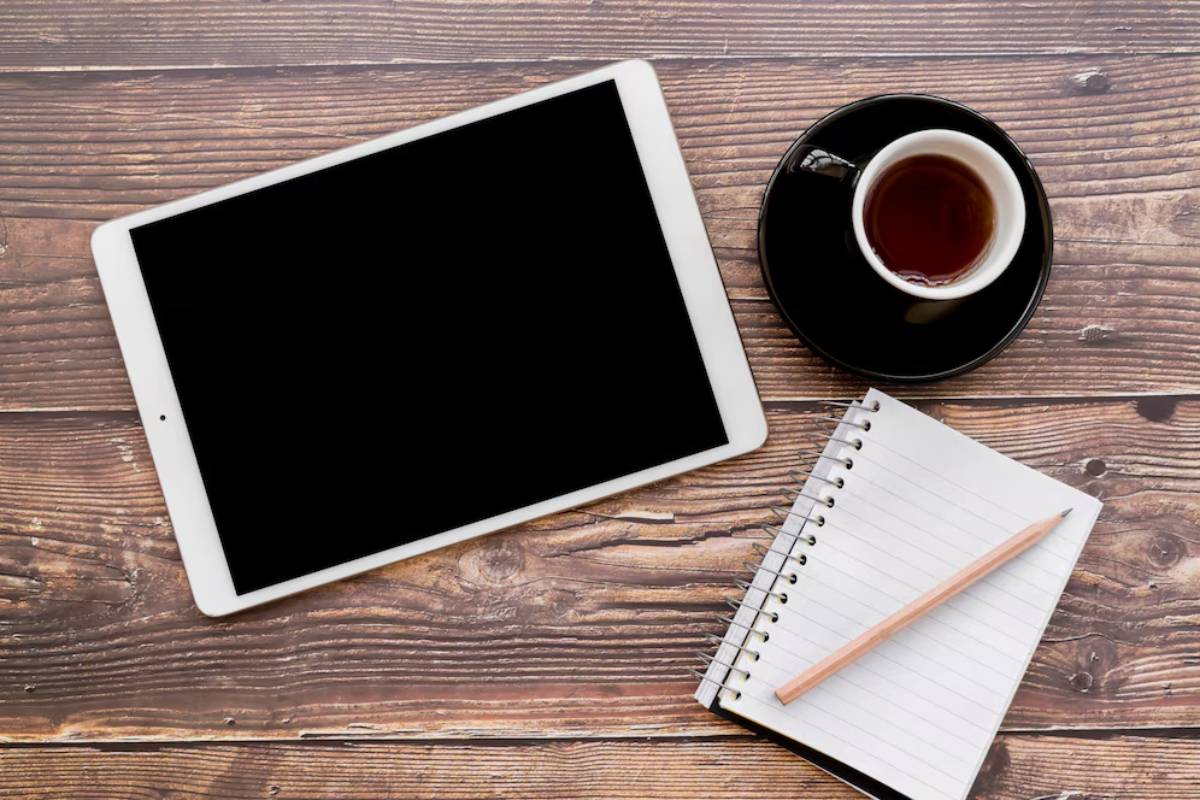 A blank tablet, a cup of tea, and a notebook with a pencil on a rustic wooden table, suggesting a workspace for ideas and creativity.