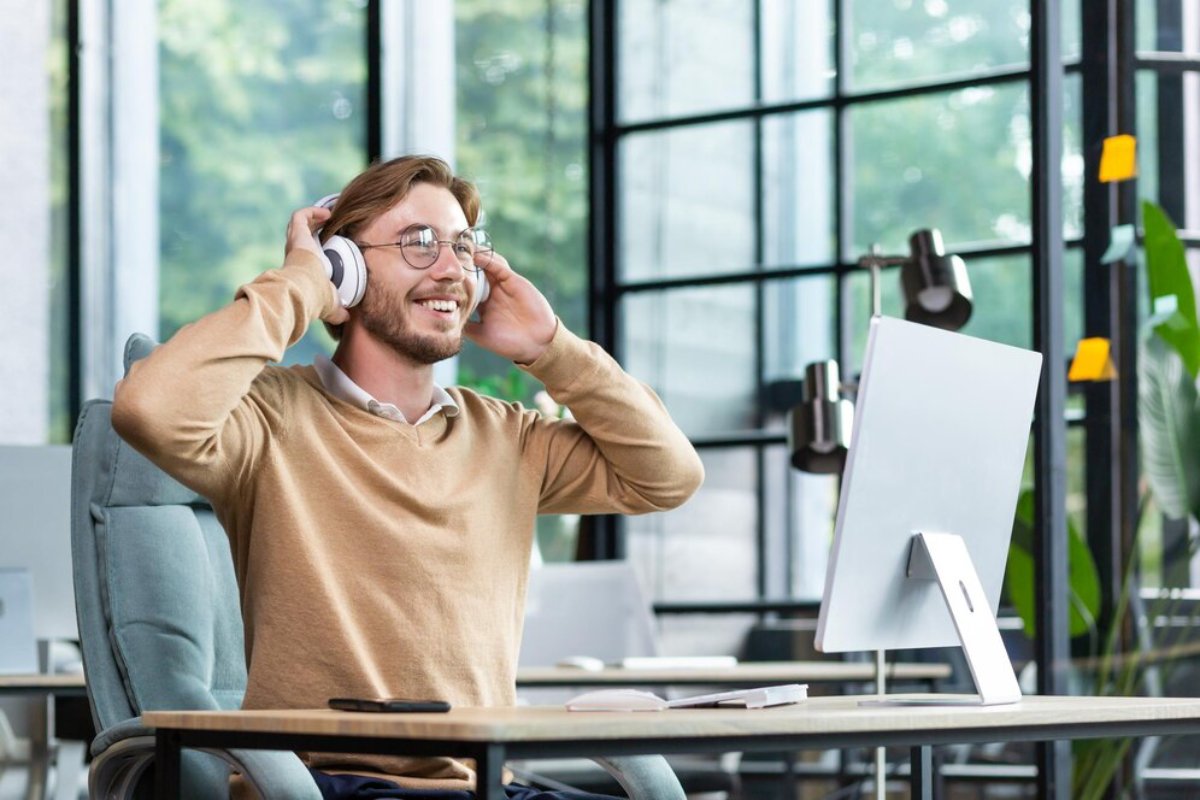 Smiling man with headphones enjoying music at a modern office desk, surrounded by plants and large windows.