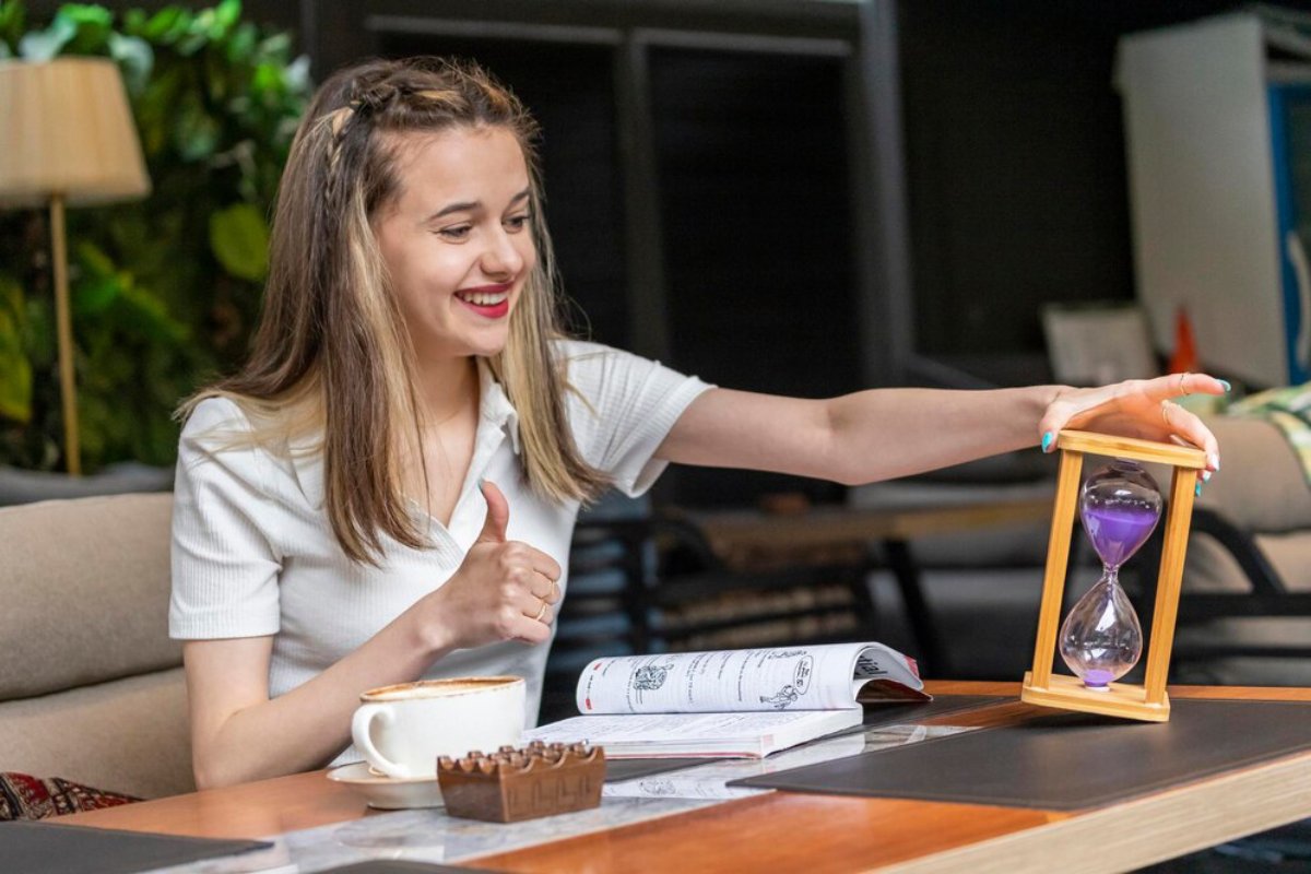 Young woman smiling and giving a thumbs up while holding an hourglass at a cafe table with coffee and a book.