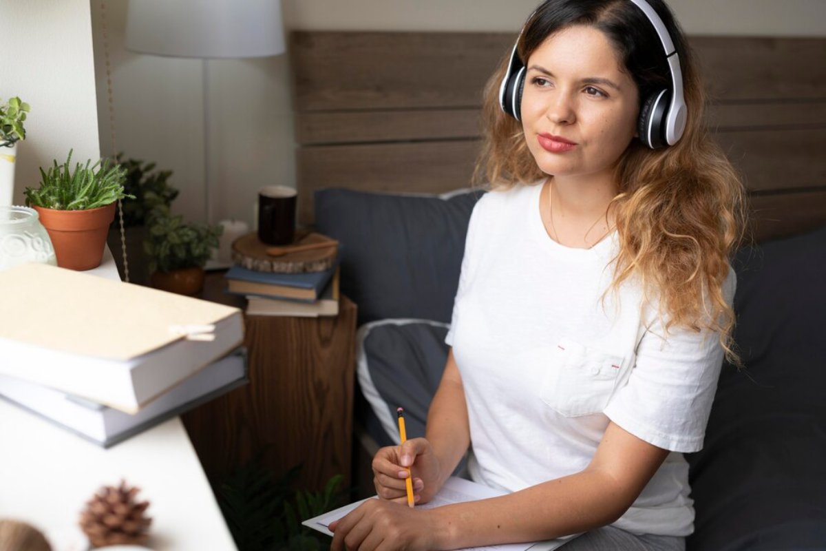 Woman wearing headphones while writing in a notebook, sitting at a cozy home study area with plants and books.