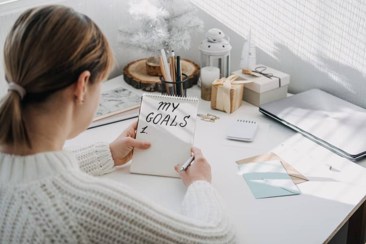 Woman writes in a notebook labeled MY GOALS at a bright, tidy workstation with festive decorations.