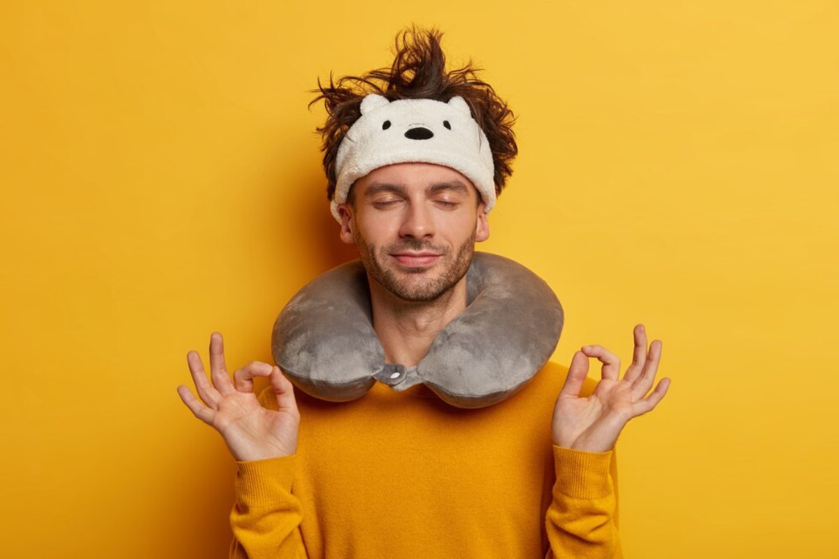 Man in yellow sweater and travel pillow, meditating against a yellow background.