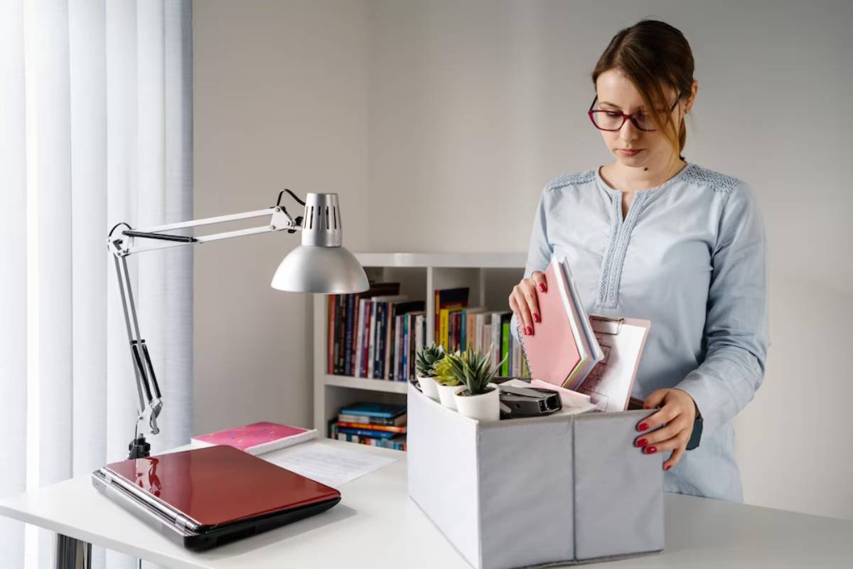 Woman packing books and supplies into a box at a desk with a laptop and lamp.