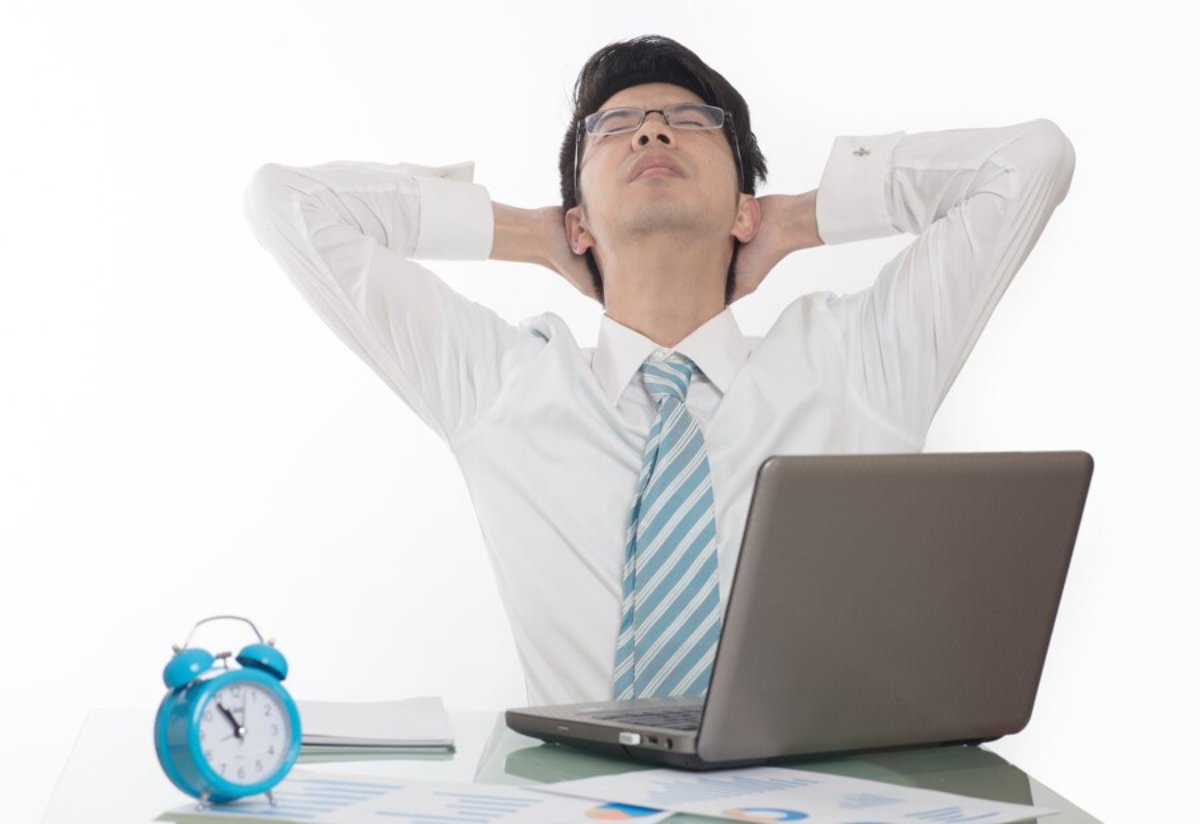 Man in white shirt and tie relaxing in his office chair with hands behind head, laptop and alarm clock on desk.