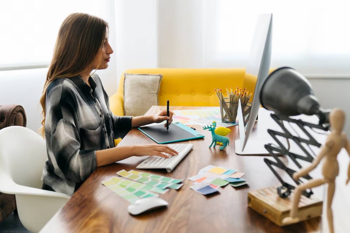 Woman using a graphics tablet at a creative workspace with a laptop, color swatches, and desk accessories.