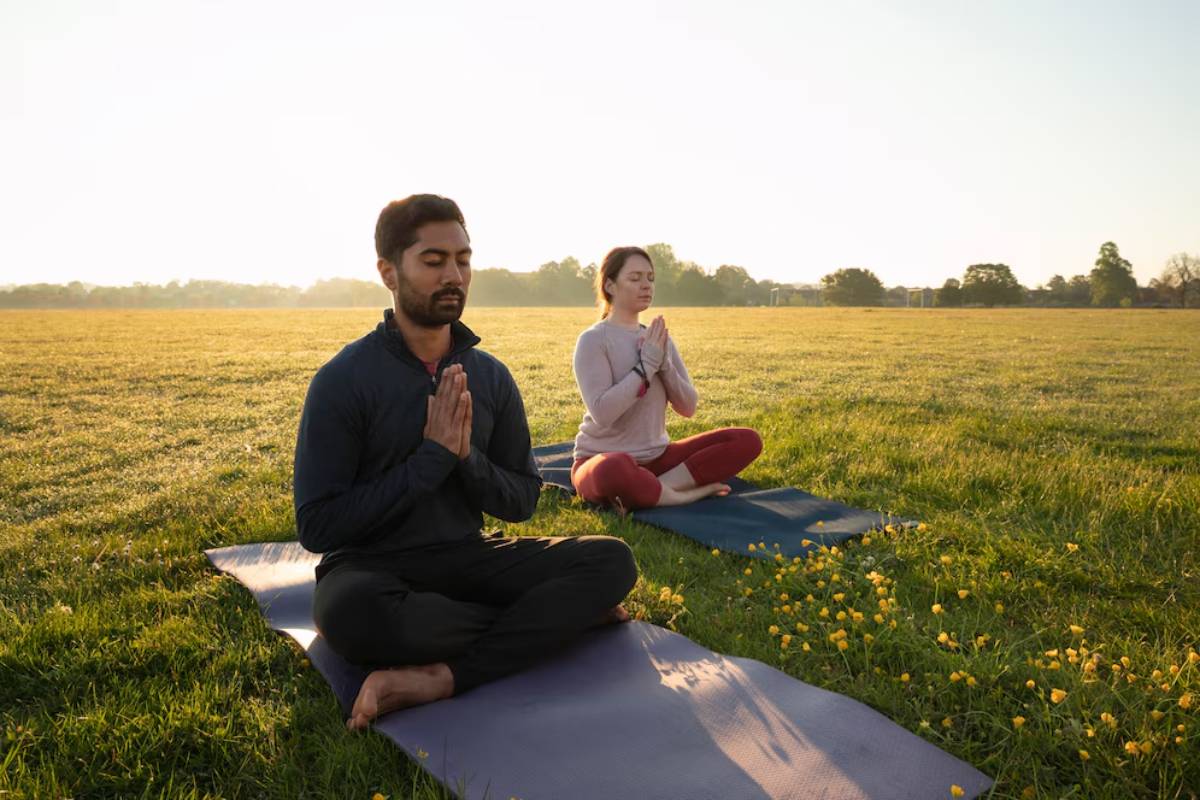Two people practicing yoga in a sunlit field, sitting on mats surrounded by green grass and yellow wildflowers.