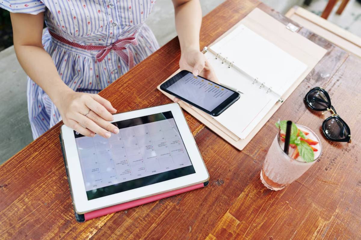 A person sits at a wooden table, using a tablet to view a calendar, with a smartphone and a refreshing drink beside them.