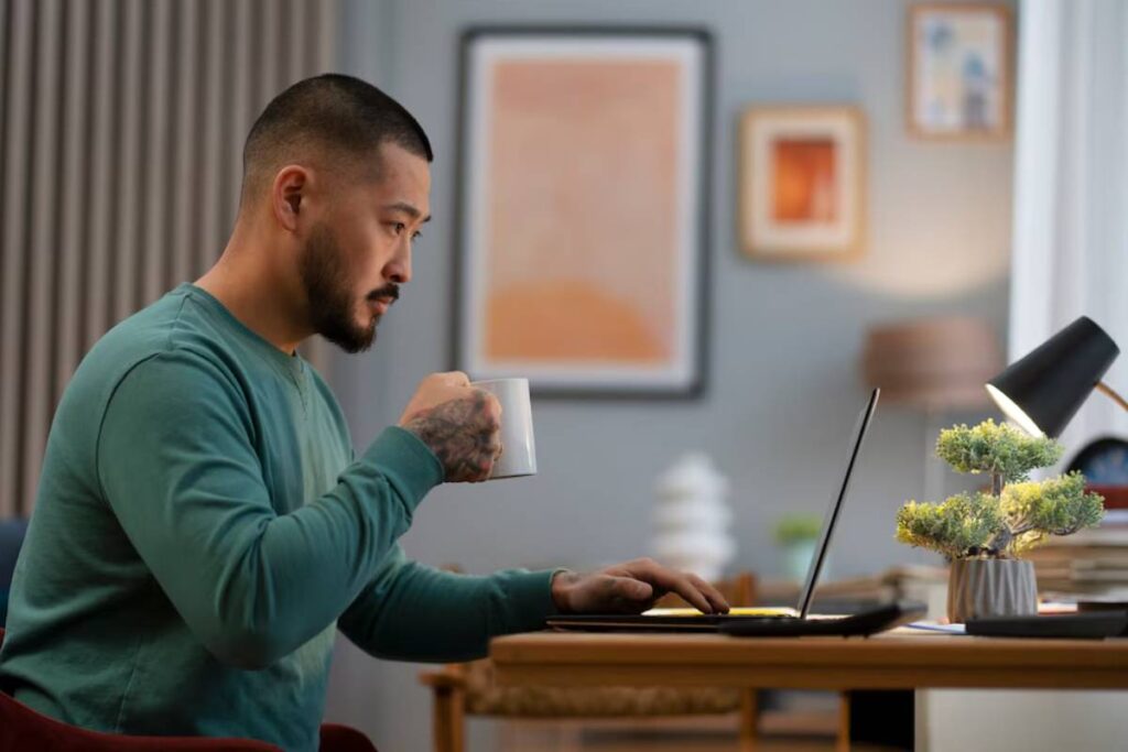 Man with tattooed arm drinking coffee and working on laptop in a home office.