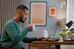 Man with tattooed arm drinking coffee and working on laptop in a home office.