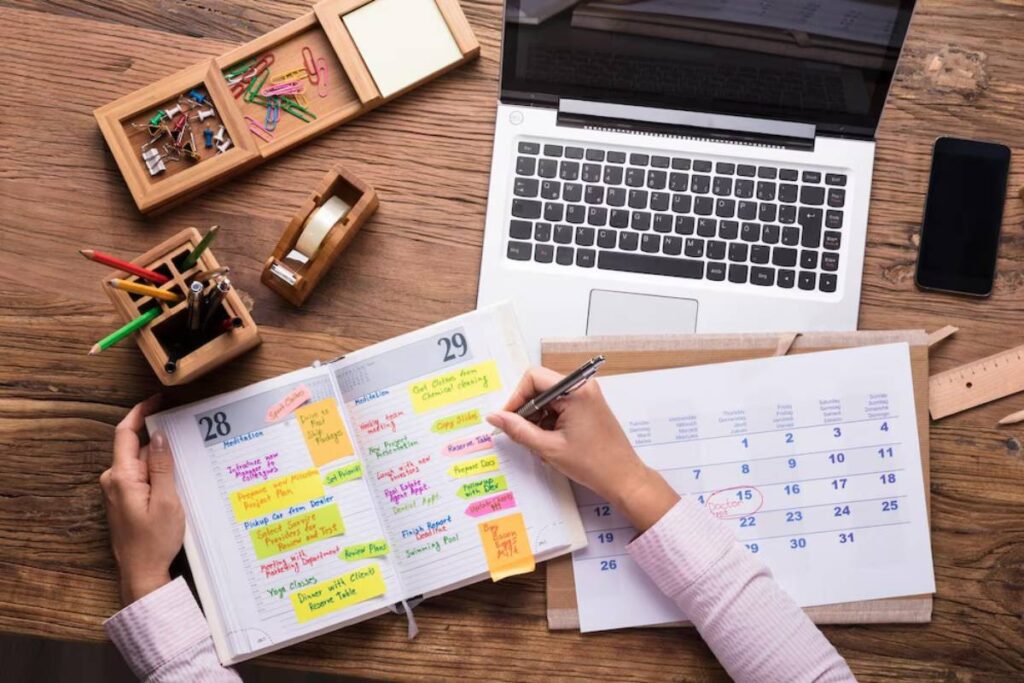 Person planning with a colorful sticky-noted planner, surrounded by office supplies and a laptop on a wooden desk.