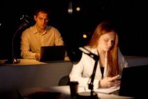Two people working on laptops in a dark office, illuminated by desk lamps.