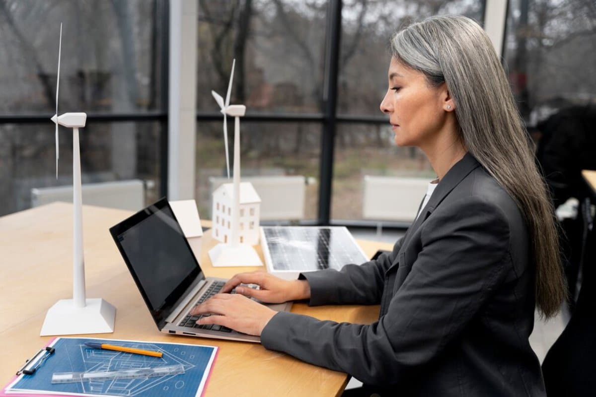 A professional woman works on a laptop surrounded by models of wind turbines and solar panels in a modern office.